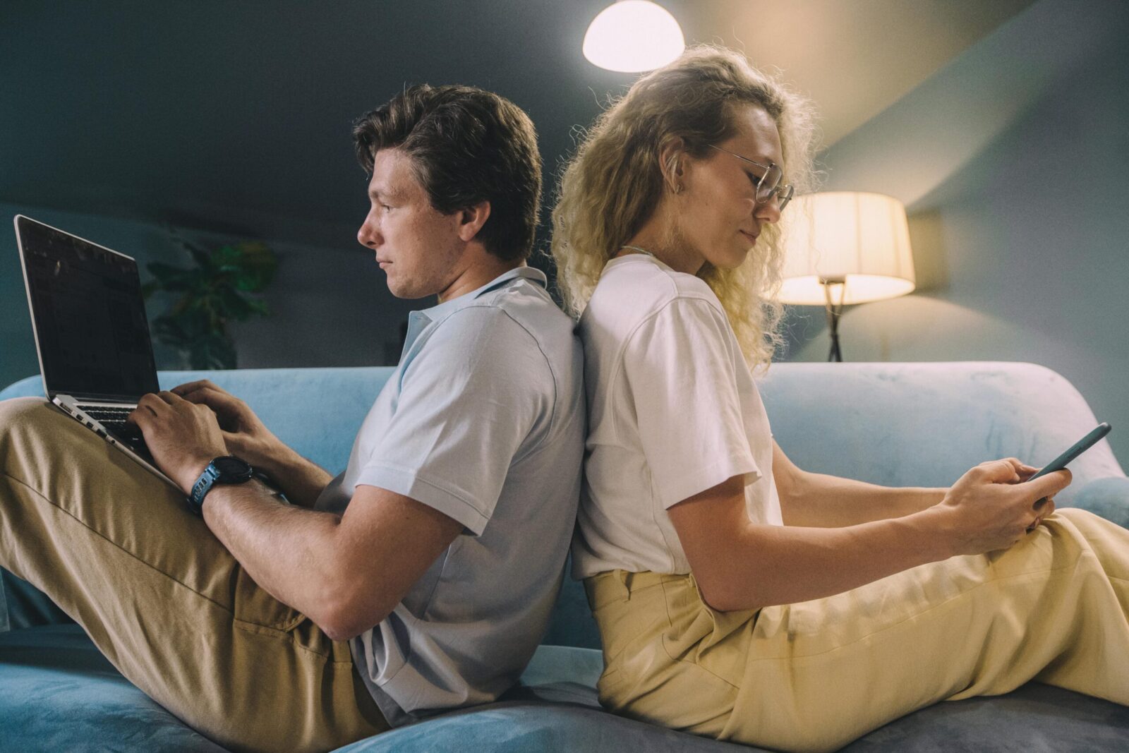 A young couple sits back-to-back, multitasking on digital devices in a cozy living room.